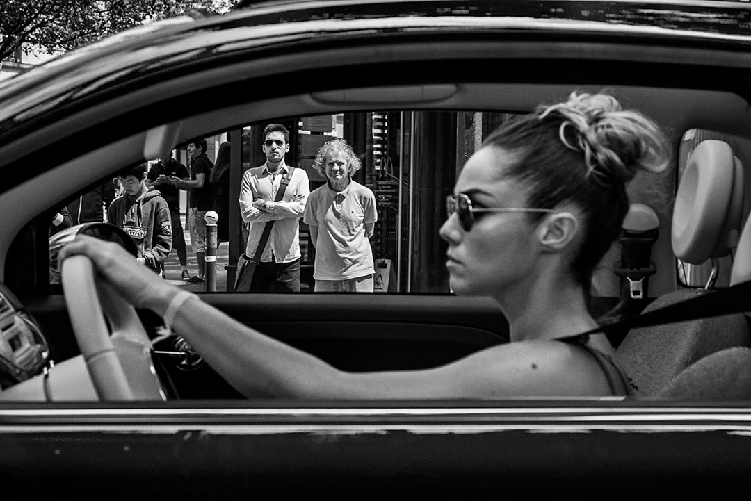 A woman driving a convertible and people on the pavement.